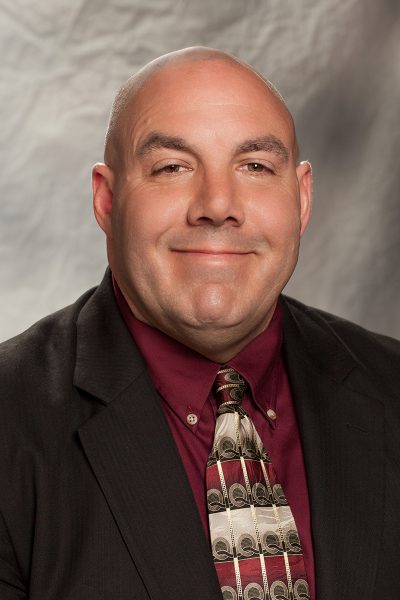 portrait photo of Chuck Pasquarell, a white man wearing a brown suit jacket with a maroon shirt and tie underneath