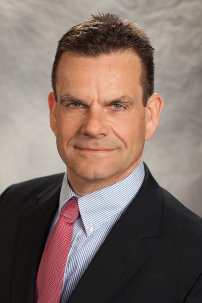 portrait photo of Thomas Uncher, a man wearing a black suit, a blue and white striped button down shirt, and a red tie
