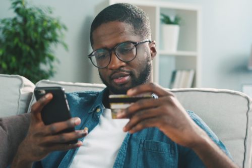 adult male wearing round black glasses holding a credit card in one hand and his cellphone in the other making an online purchase