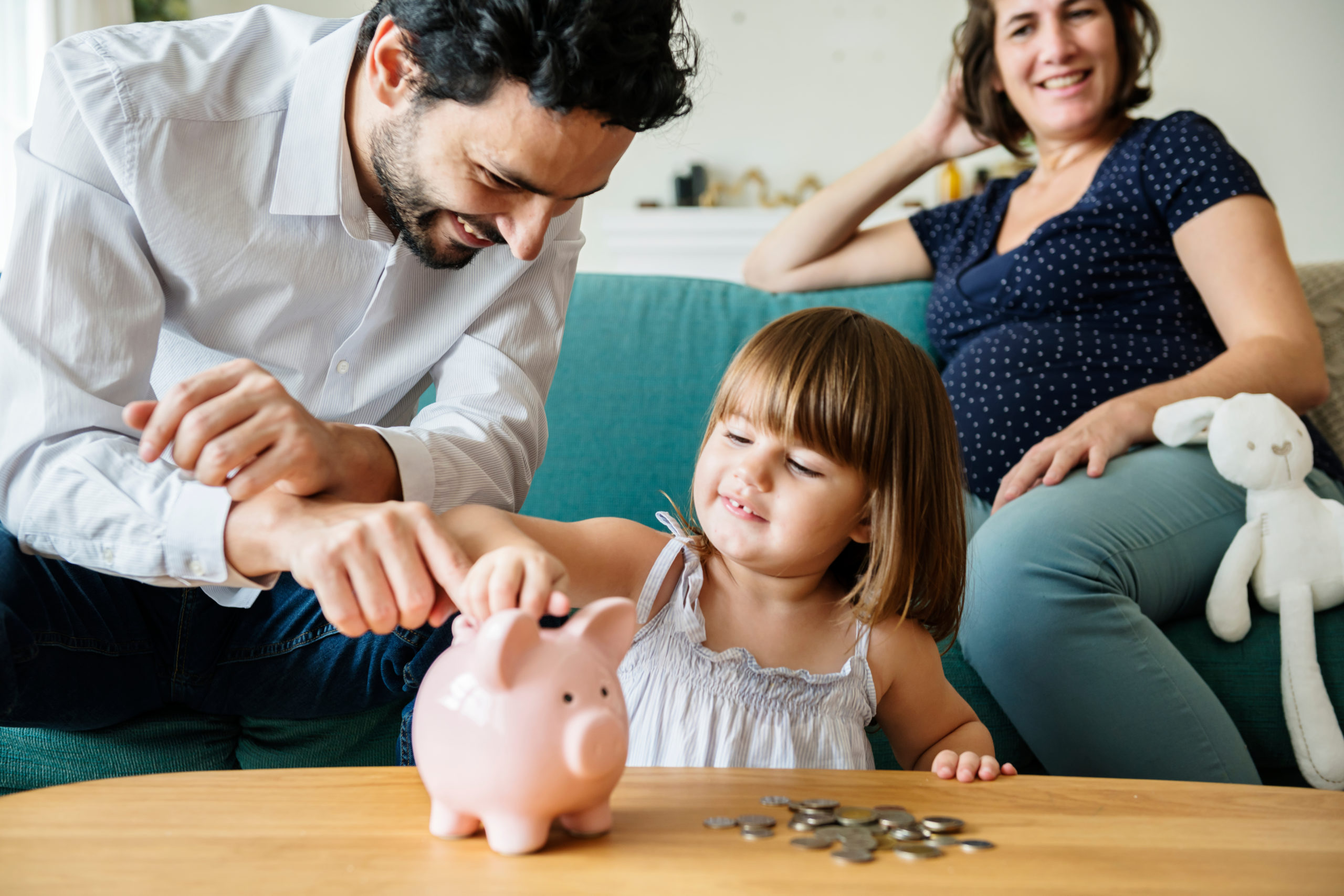 father helping young daughter put change in light pink piggy bank on a light brown wood table