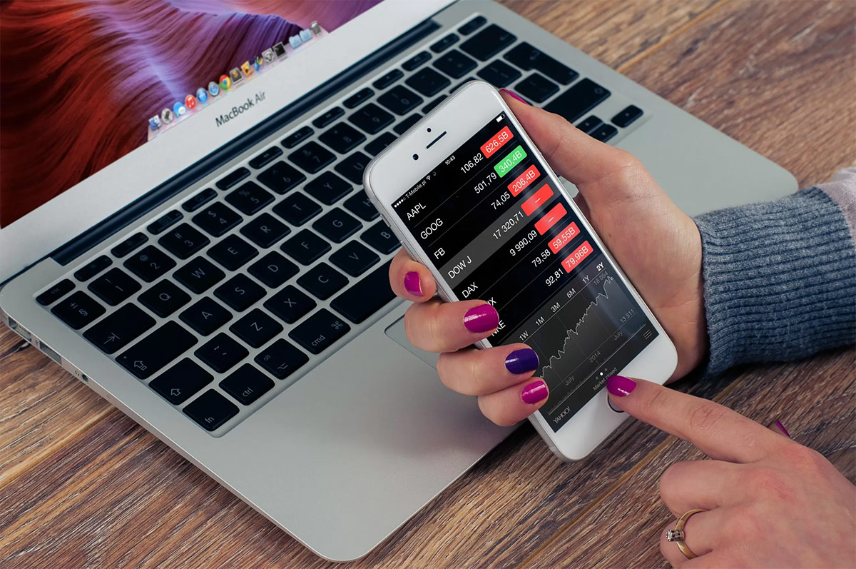 woman with pink and purple painted fingernails holding a white iPhone looking at the stock market rates