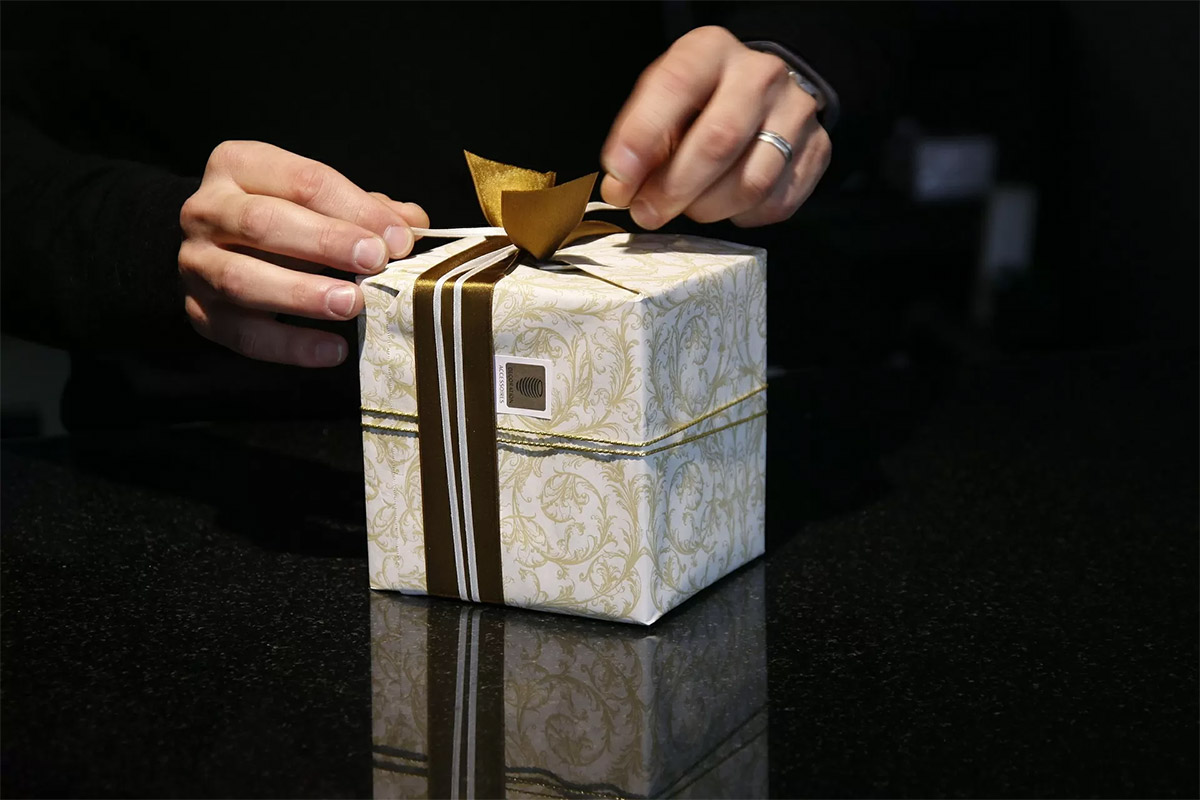 adult man wearing a black sweater tying a gold bow on a holiday gift wrapped in gold and white wrapping paper