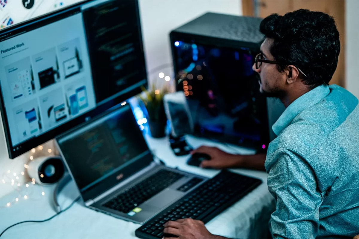 adult man with black hair and glasses wearing a blue collared shirt working on a silver laptop with a large computer monitor behind it