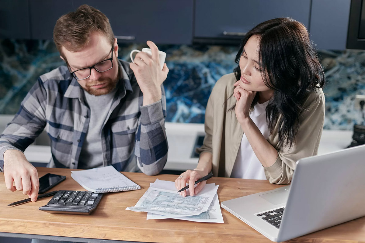 adult man holding a white coffee mug sitting next to an adult woman looking for financial paperwork next to a black calculator and silver laptop
