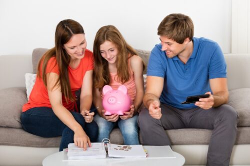 young teen holding a pink piggy bank sitting on a gray couch between two adults look at a planner