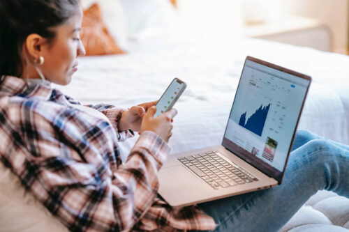 female student in blue and white plaid shirt looking at smartphone in her hands with a silver laptop on her lap