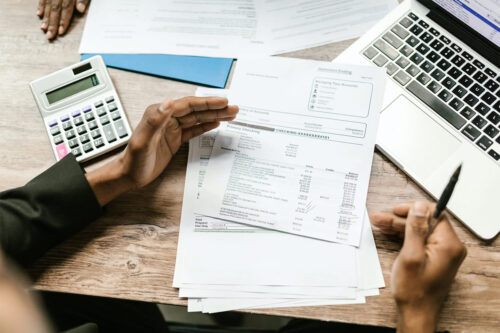 overhead shot of three hands resting on a brown, wooden desk with white financial documents near a silver laptop, blue folder, and silver calculator