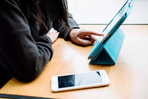 woman in black jacket sitting at brown, wooden desk accessing a mobile banking app on iPad in a sky-blue case with a white iPhone next to it