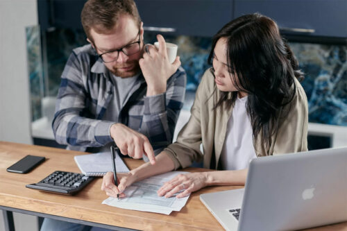 man and woman sitting at tan desk calculating their DTI ratio using white notepad, silver laptop, a black iPhone, and calculator