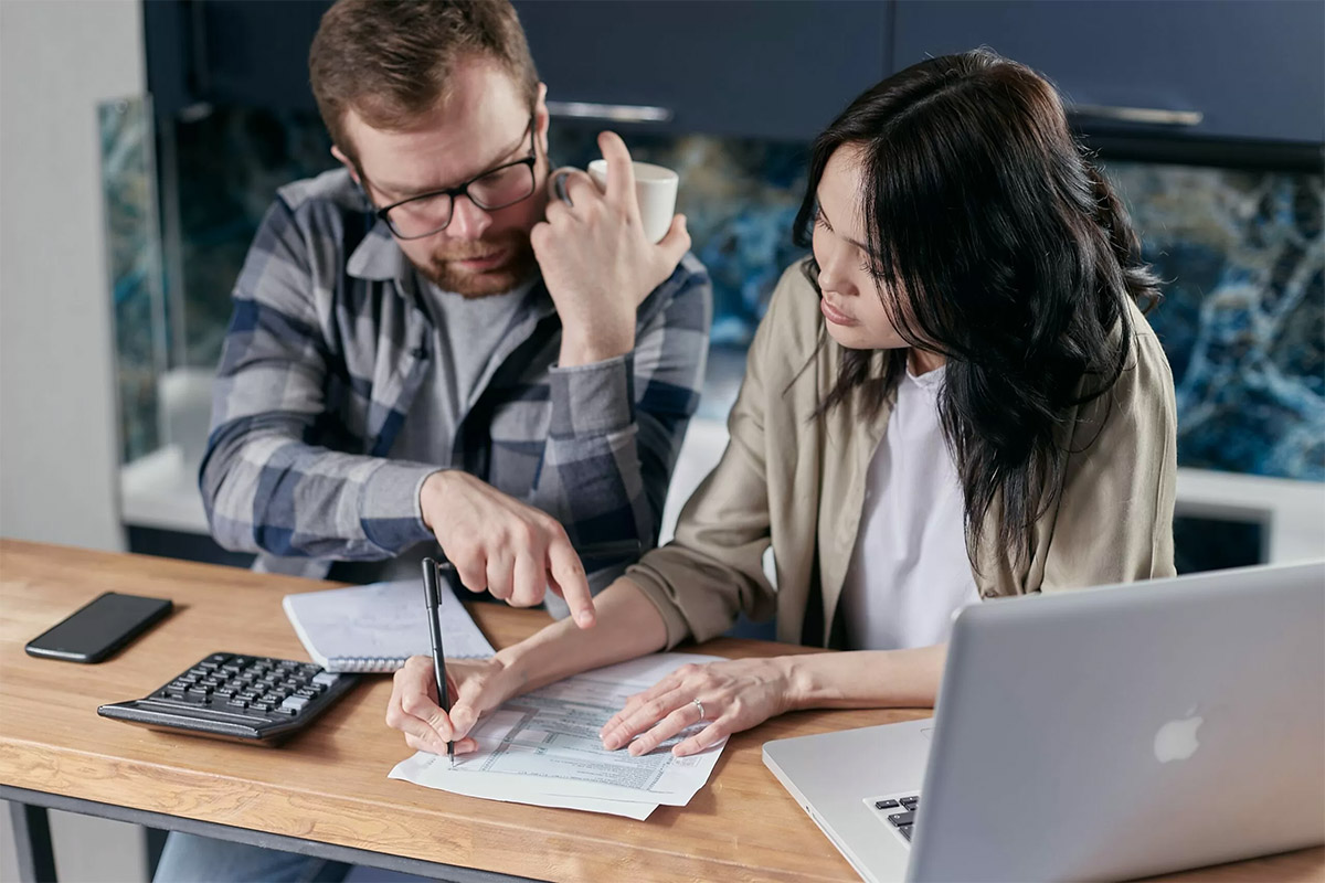 man and woman sitting at tan desk calculating their DTI ratio using white notepad, silver laptop, a black iPhone, and calculator
