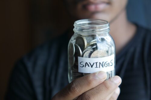 man wearing a black shirt holding a glass mason jar filled with coins that has been labeled “savings”
