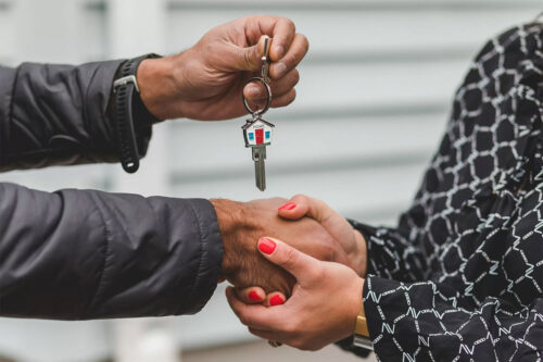 woman with red nail polish on her fingers wearing a black shirt with a white pattern shaking the hands of a man wearing a black coat, holding house keys