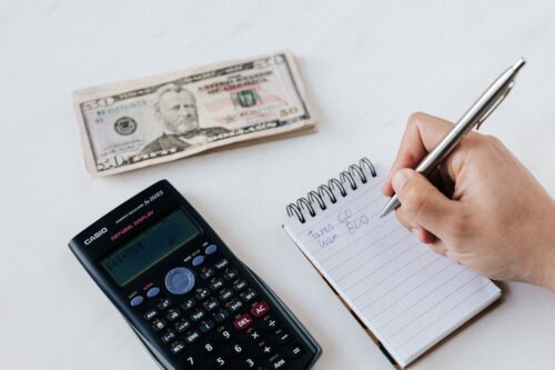 adult hand holding a silver pen writing finances in a small notepad next to a black calculator and money