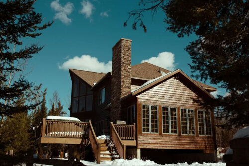 brown wooden house with snow on the deck and a stone chimney in need of maintenance