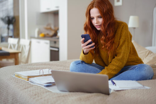 Female Sitting on Bed with Laptop Computer, Paperwork and Checking Cell Phone while Working From Home