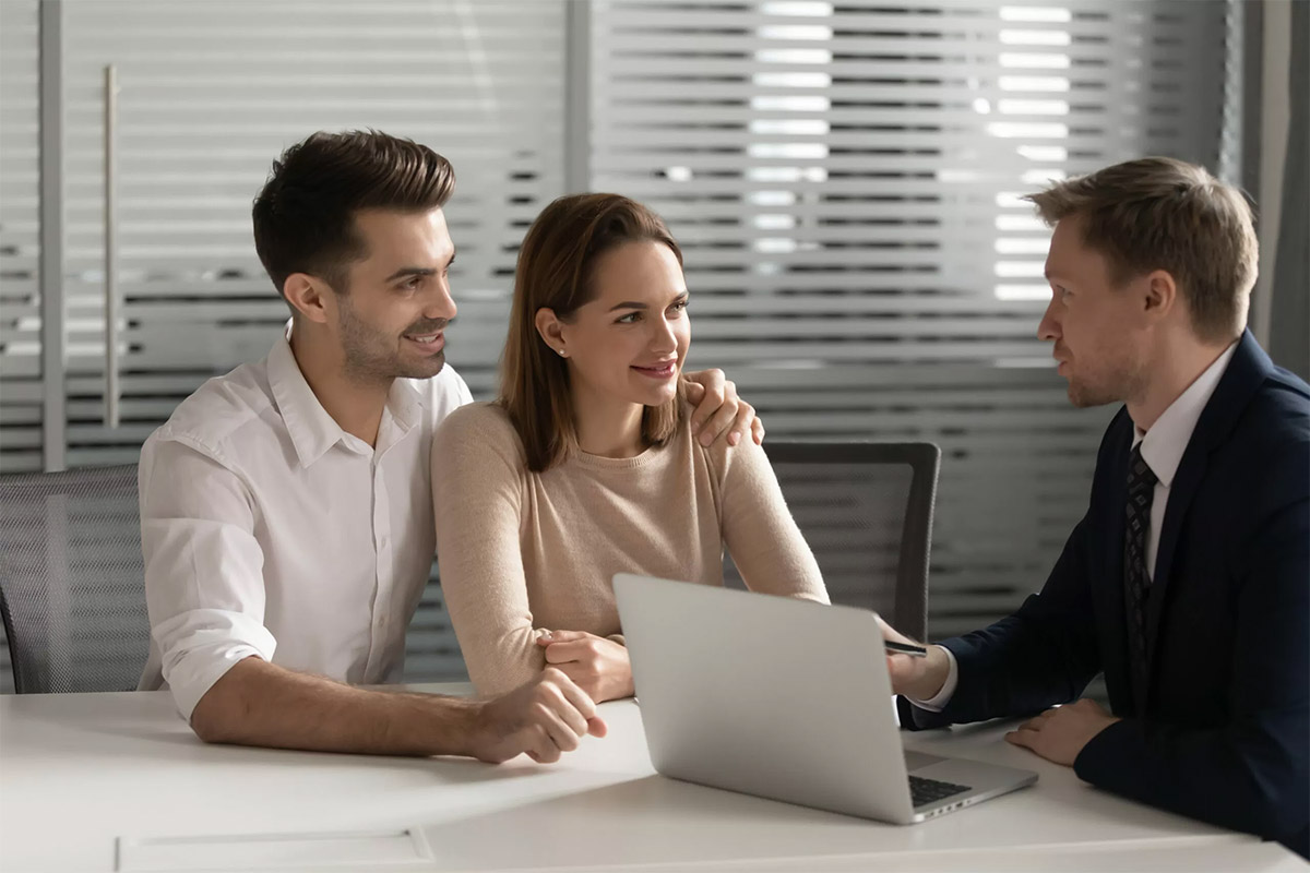 Happy millennial couple talk with male real estate agent or broker in office buying first house together, smiling young husband and wife speak visit banker think of taking loan or mortgage from bank