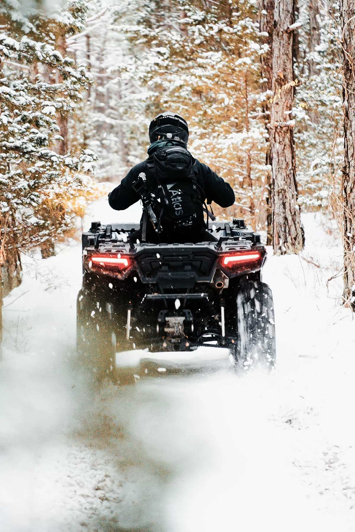 person dressed all in black with a black helmet riding an ATV on a snowy, tree-lined trail