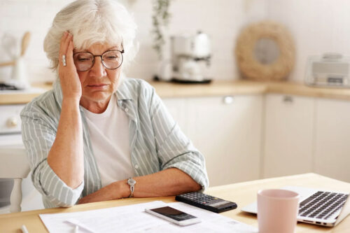 elder woman holds hand to head at kitchen table as she looks at finances