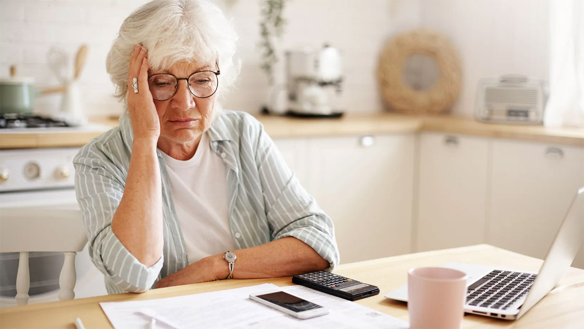 elder woman holds hand to head at kitchen table as she looks at finances