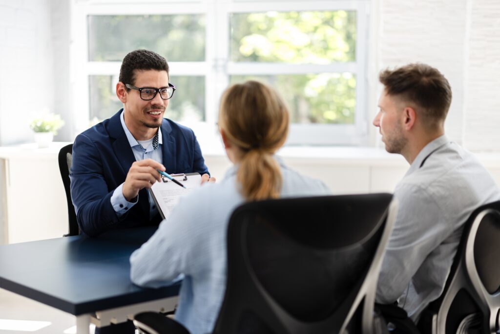 Mortgage loan officer sitting at a desk, holding a pen and clipboard explaining to two people seated in chairs the mortgage process.