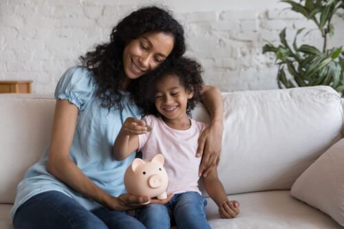 mother and daughter sitting on a white couch while the daughter is putting money into a piggy bank
