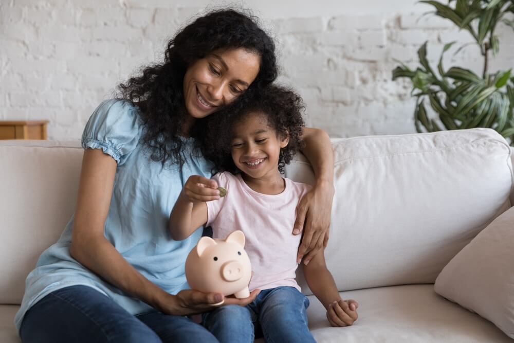 mother and daughter sitting on a white couch while the daughter is putting money into a piggy bank