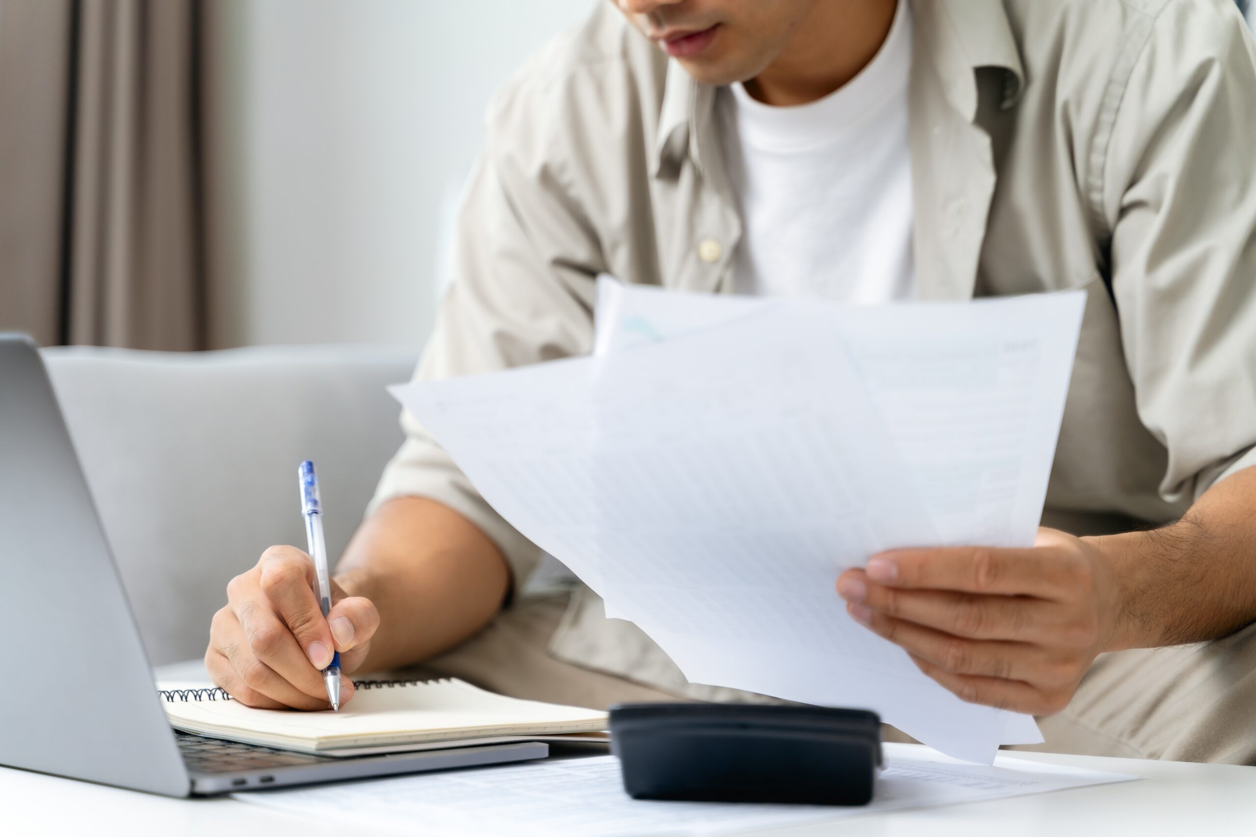 man calculating finances while sifting through papers on the couch writing down numbers on a pad of paper